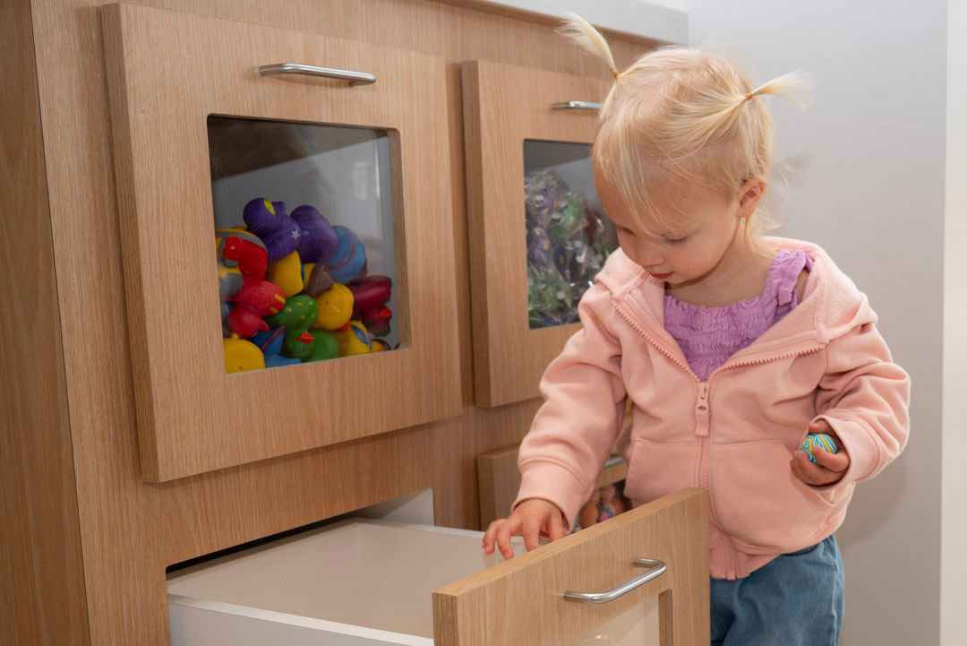A young child standing next to a kitchen cabinet with colorful balls inside.