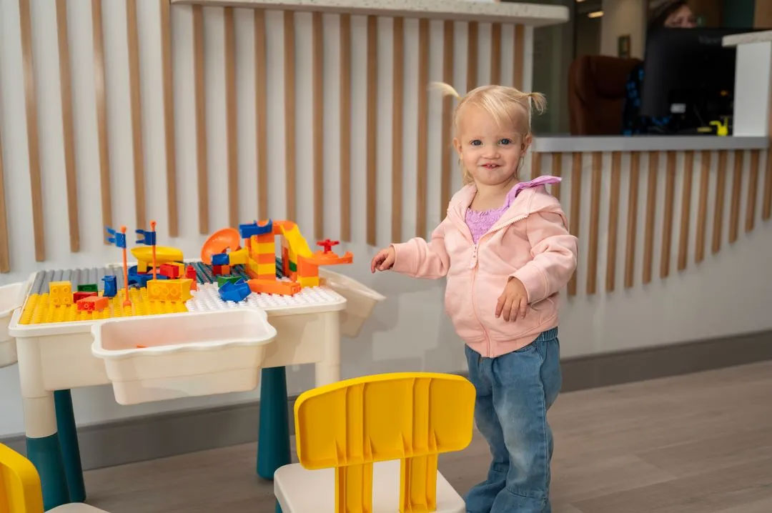 A young child standing next to a colorful play table with toys, wearing a pink jacket and blue jeans.