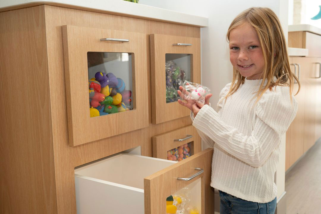 A young girl holding a toy while standing next to a kitchen cabinet filled with colorful balls.