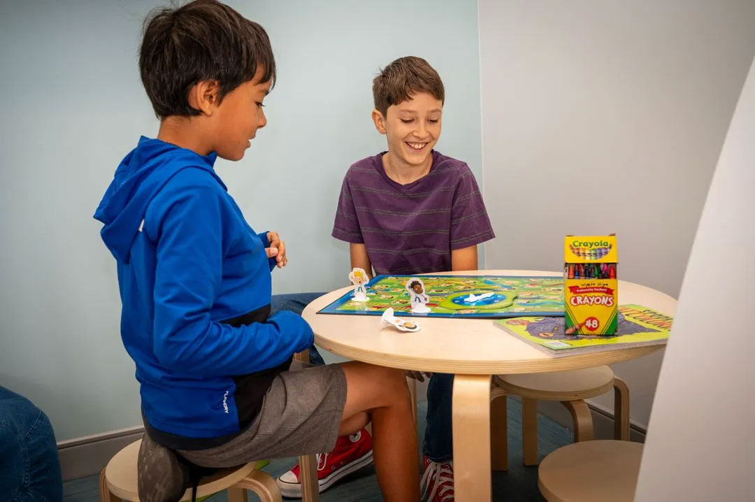 Two young boys sitting at a table with a toy set in front of them, smiling and engaged in play.
