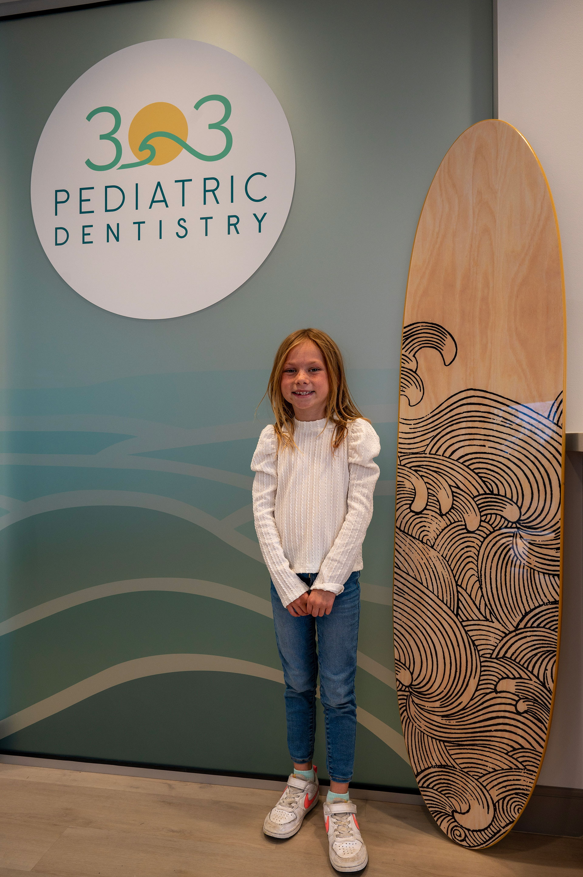 A young girl standing next to a surfboard with the logo '303 Pediatric Dentistry' on a wall.