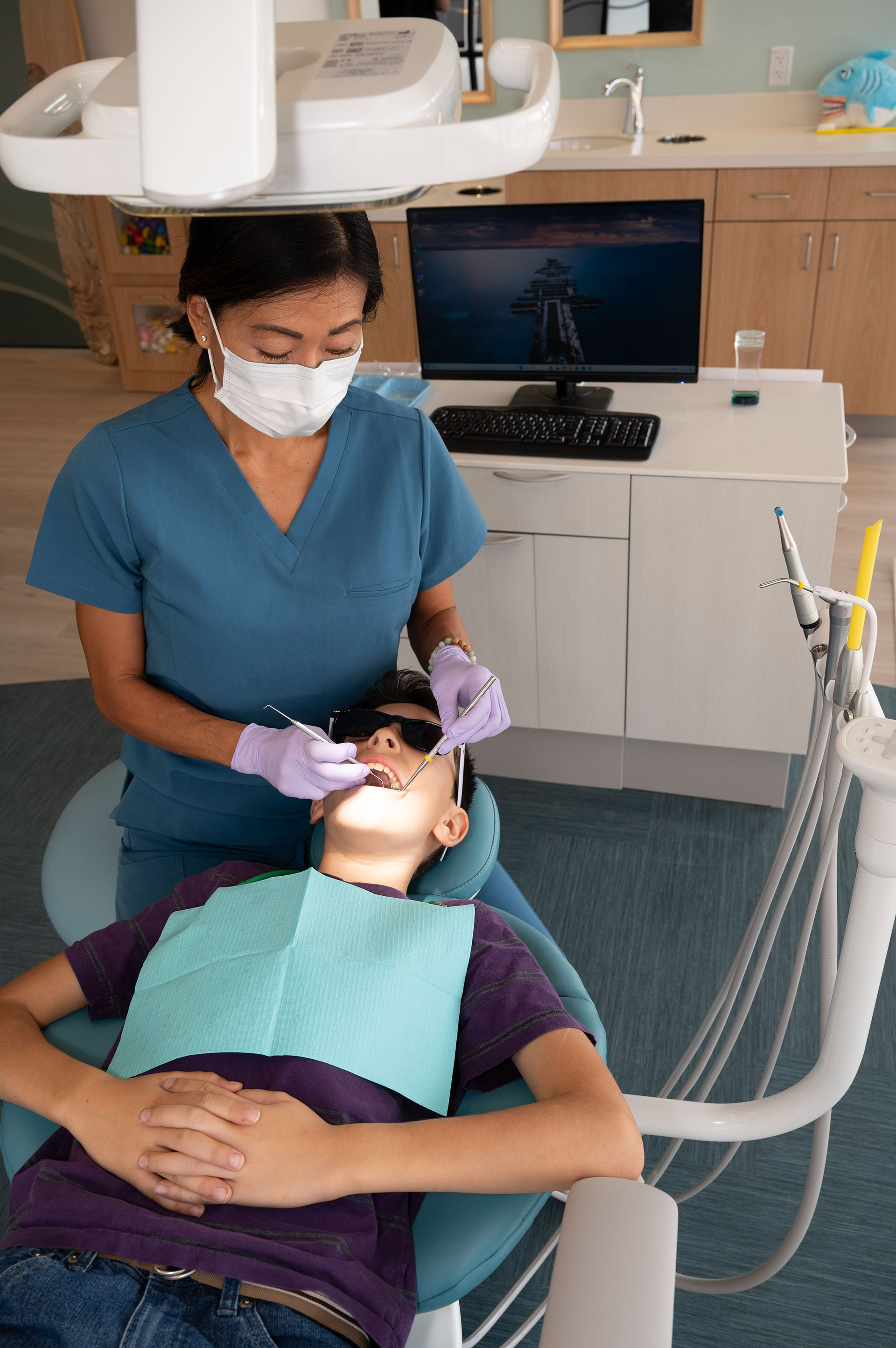 A dental hygienist wearing a face mask is assisting a patient with a dental appointment, using a dental chair with advanced technology for examination.