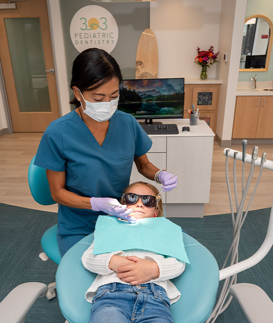 A dental hygienist in a face mask is attending to a patient who is seated in a dental chair with their mouth open, receiving dental care.
