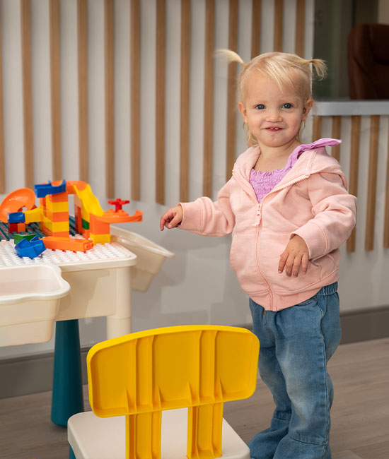 A young child standing next to a toy kitchen set with a smile on their face.