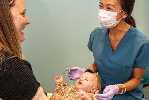 The image shows a woman in a medical gown and mask examining a baby with a stethoscope while another person watches.