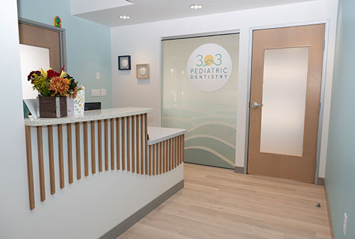 The image shows an interior view of a dental office waiting area with a reception desk, a window displaying a logo and text, a counter with a sign, and a door with a glass panel.